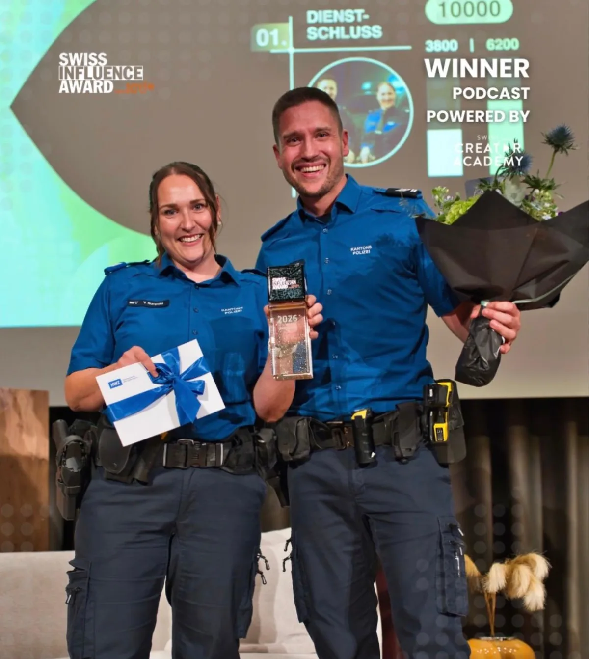 Zwei lächelnde Personen in blauer Polizeiuniform halten auf einer Bühne eine Auszeichnung und Blumen in Händen.