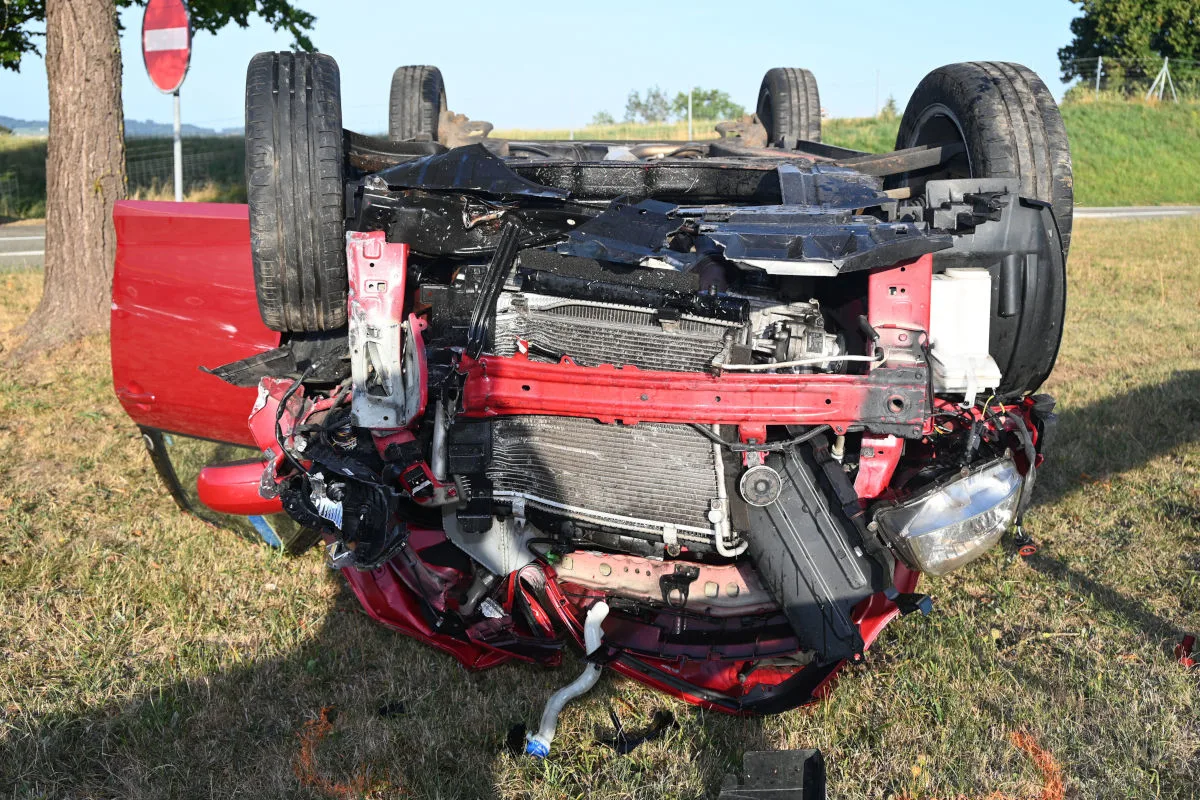 Ein rotes Auto liegt auf dem Dach mit sichtbaren Schäden an der Karosserie. Der Unfall ereignete sich auf einer Wiese neben einer Straße.