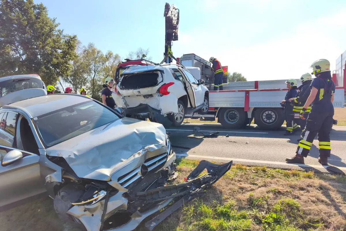 Ein silbernes Auto mit Frontschaden steht auf einer Straße. Feuerwehrleute verladen ein beschädigtes weißes Auto auf einen Lkw.