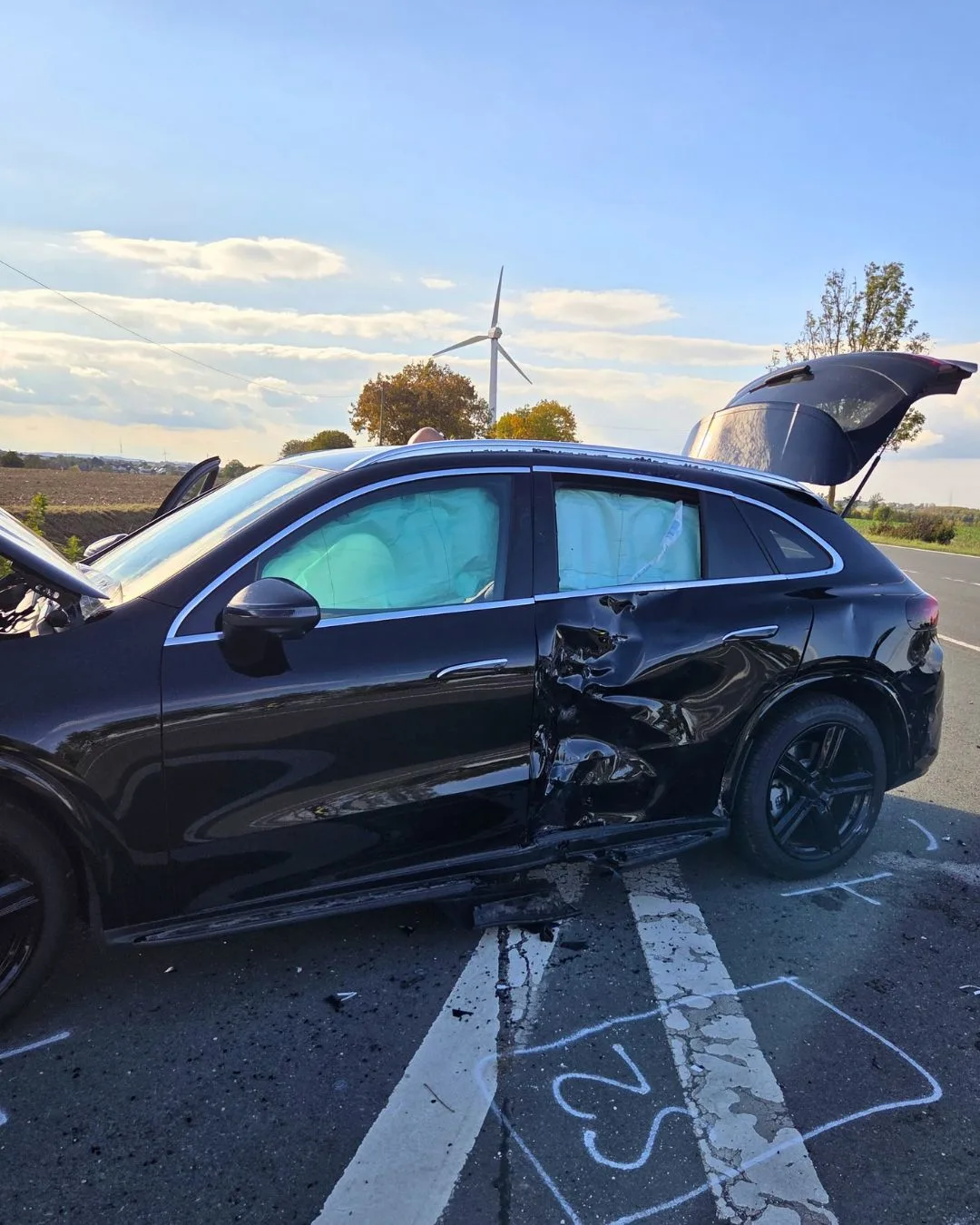 Ein schwarzes Auto mit erheblichem Unfallschaden an der Fahrerseite steht auf einer Landstraße. Im Hintergrund sind Windräder und Bäume zu sehen.