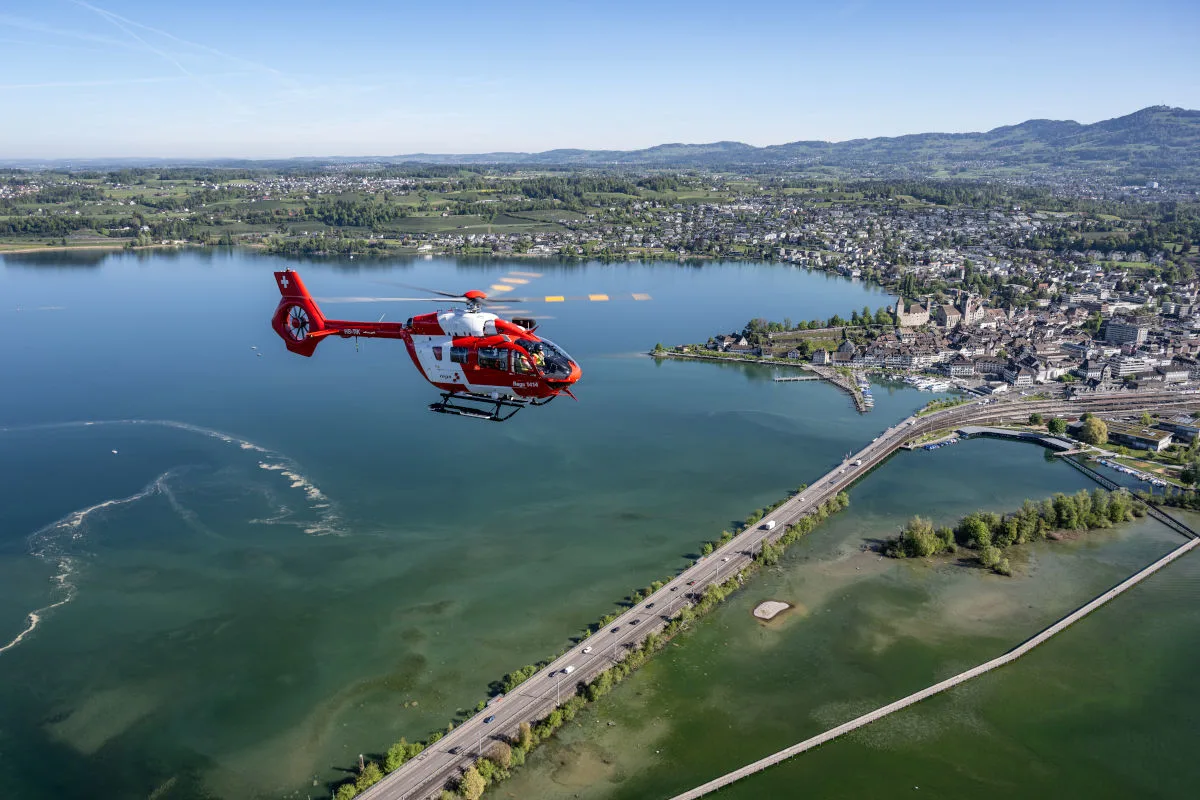 Ein roter Rettungshubschrauber fliegt über einen weitläufigen See und eine Stadtlandschaft mit Bergen im Hintergrund.