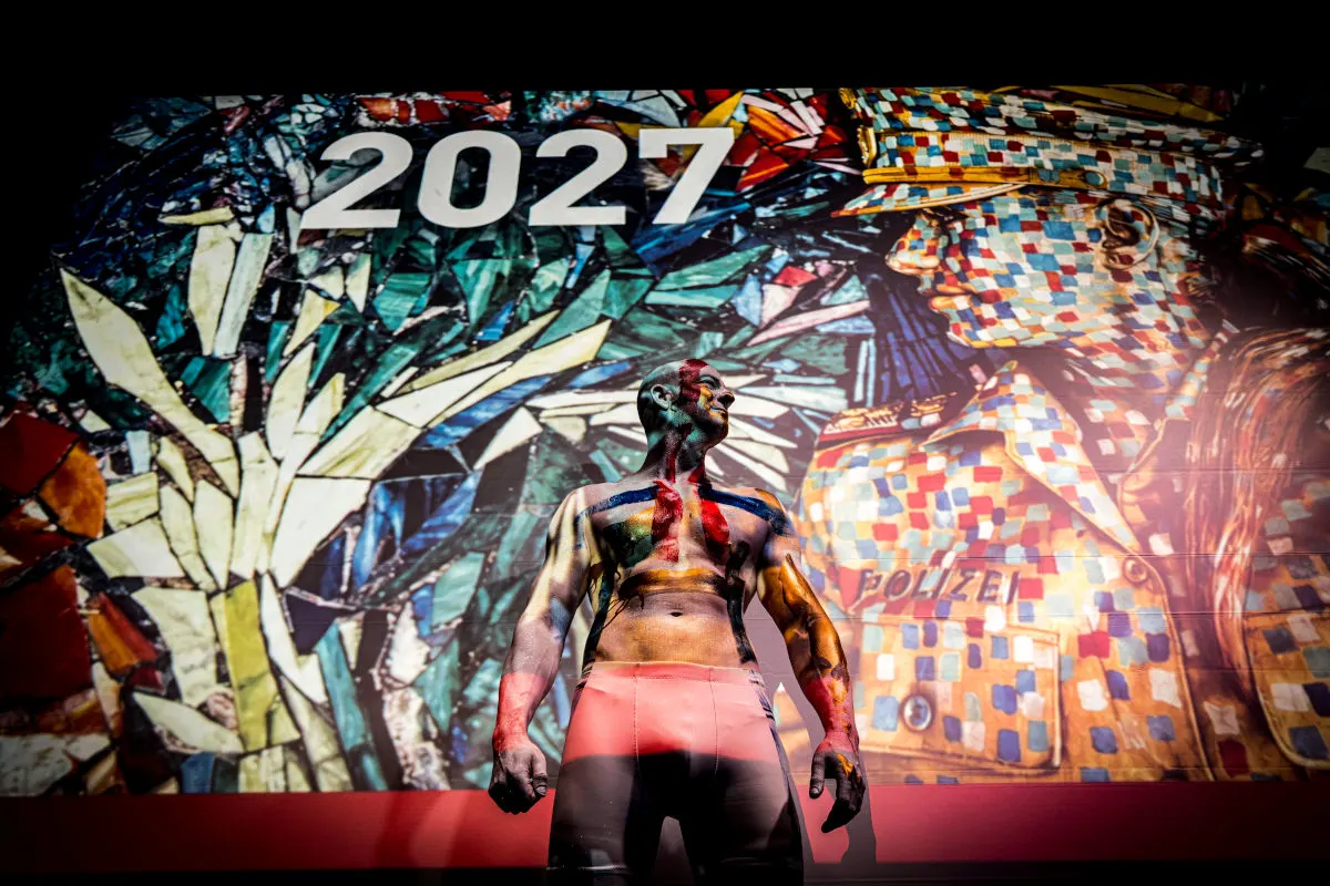 Ein Mann mit bemaltem Körper steht vor einer projizierten, bunten Kunstinstallation, die eine Polizistfigur und die Zahl 2027 zeigt.