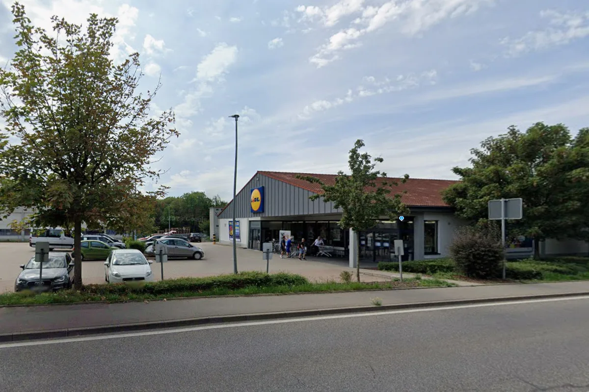 Ein Supermarkt mit einem Parkplatz im Vordergrund, einige Autos sind geparkt und Personen betreten das Gebäude.