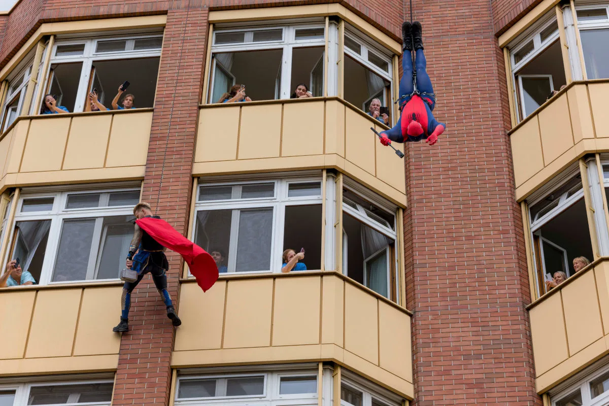 Zwei Personen in Superheldenkostümen, darunter einer als Spiderman, klettern die Fassade eines Wohngebäudes hoch. Zuschauer beobachten gespannt aus den Fenstern.
