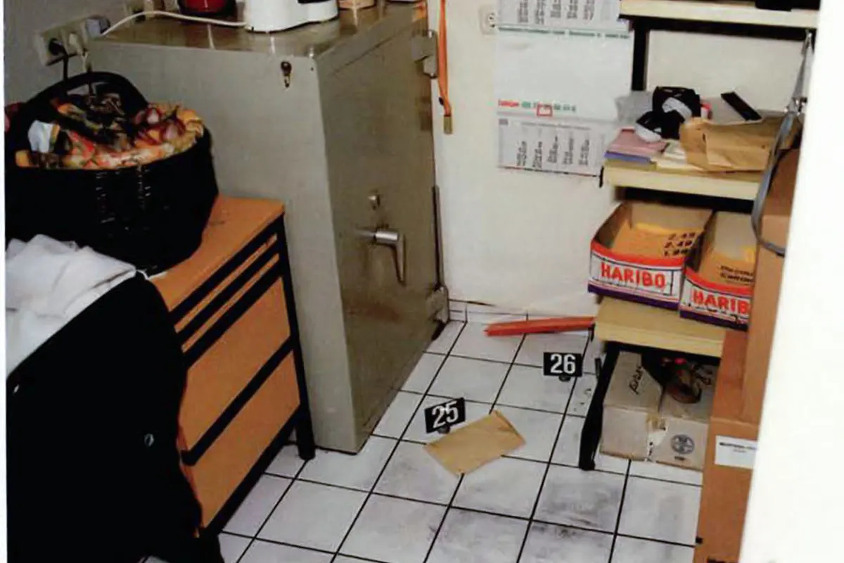 Ein Büro mit einem großen Tresor in der Ecke, mehrere markierte Beweisstücke auf dem Boden und Regale mit Kartons.