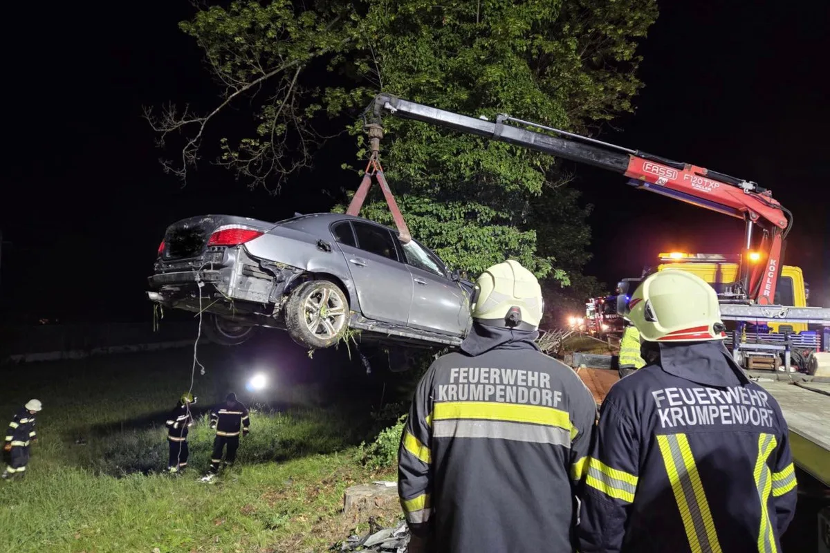 Ein Auto wird bei Nacht von einem Kran angehoben. Feuerwehrleute in Schutzkleidung beobachten die Szene. Die Umgebung ist dunkel und schlecht beleuchtet.