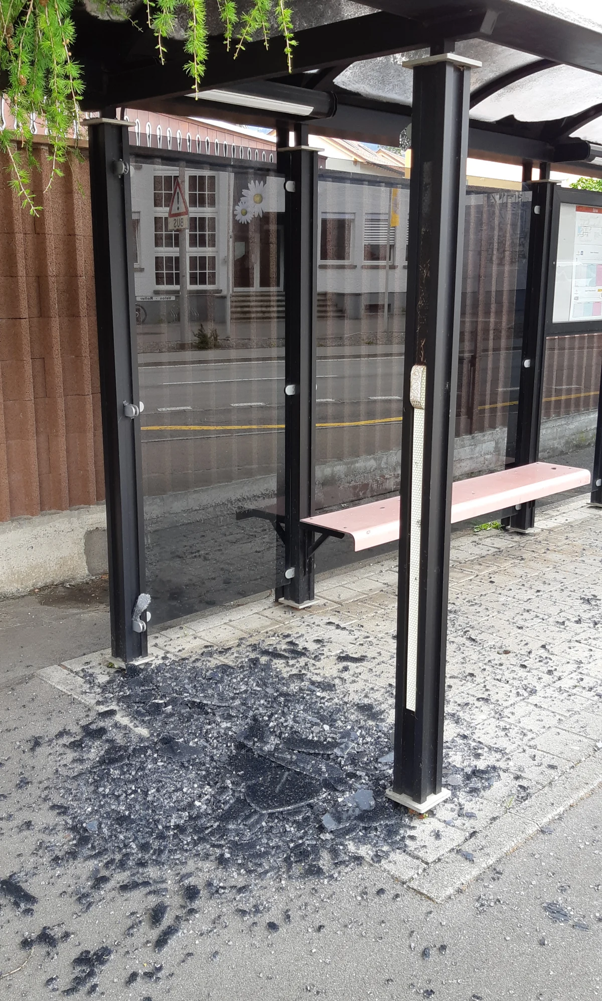 An einer Bushaltestelle ist Glas zerbrochen, Scherben liegen auf dem Boden. Die Haltestelle ist überdacht und an einer Straße gelegen.