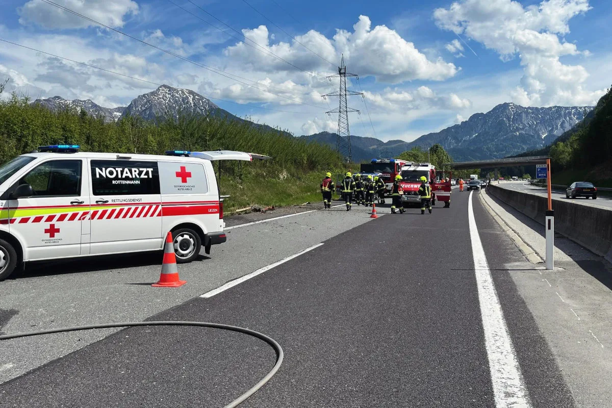 Ein Notarztwagen und Feuerwehrfahrzeuge stehen auf einer Autobahn, umgeben von Feuerwehrleuten. Im Hintergrund sind Berge und ein bewölkter Himmel zu sehen.