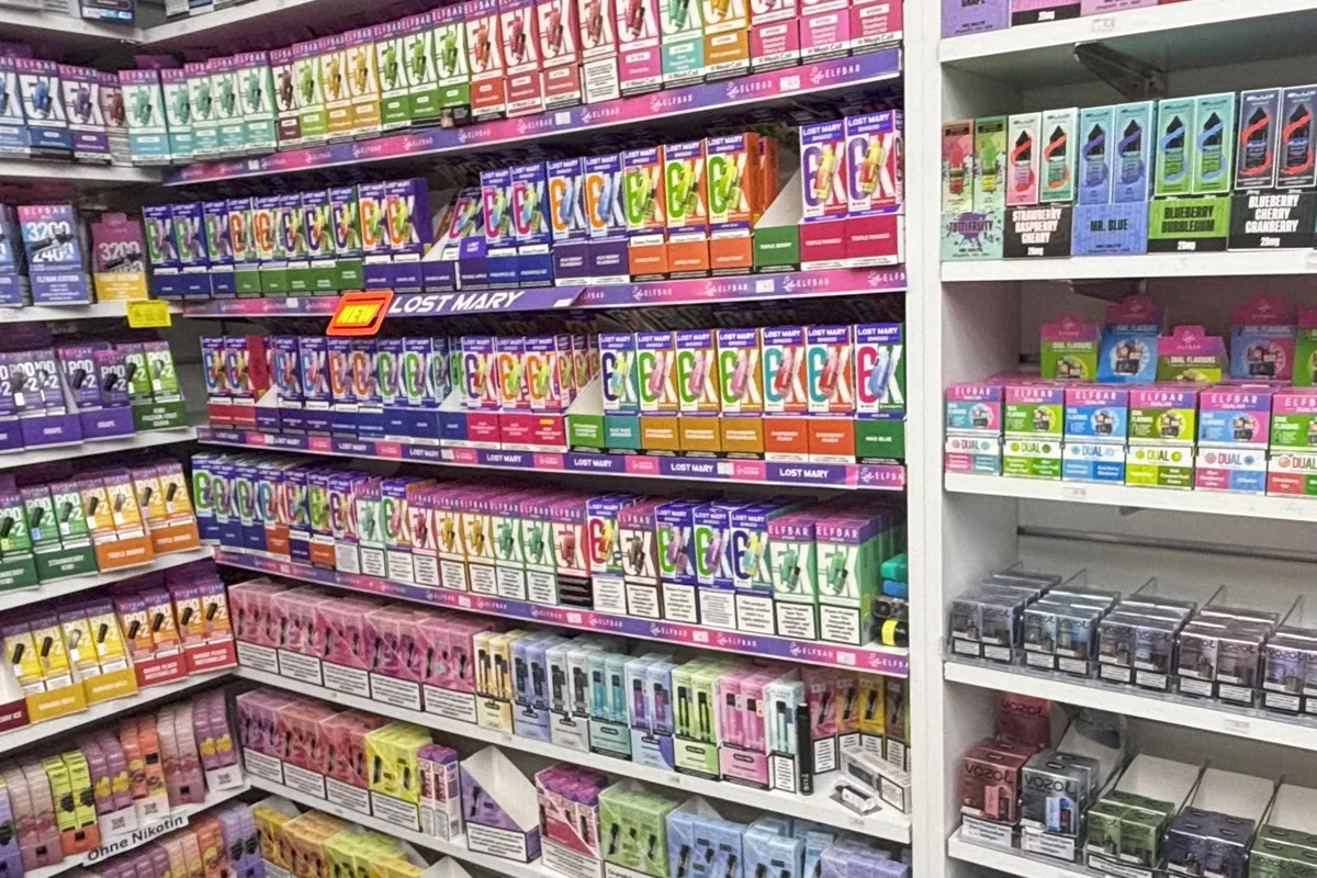Ein Ladenregal zeigt zahlreiche bunte Verpackungen von Vape-Produkten diverser Marken in verschiedenen Geschmacksrichtungen.