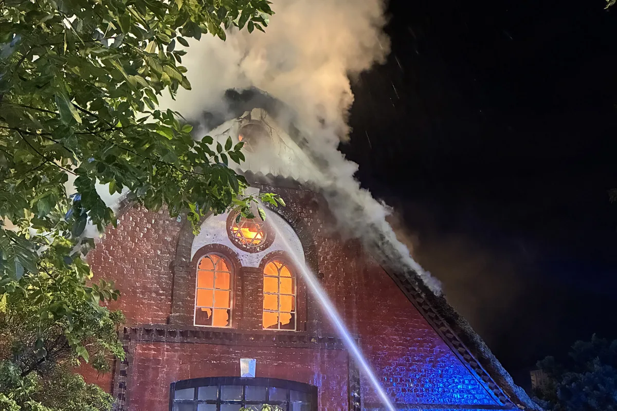 Die Feuerwehr ist dabei, ein Feuer an einem Backsteingebäude mit einem beschädigten Dach zu löschen. Rauch und Flammen sind sichtbar.