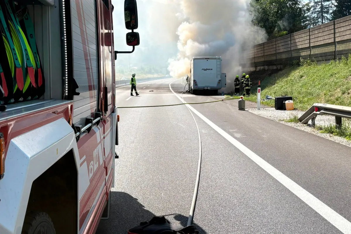 Ein Wohnmobil steht auf einer Autobahn in Flammen, während Feuerwehrleute mit dem Löschen beschäftigt sind. Ein Feuerwehrfahrzeug ist im Vordergrund sichtbar.