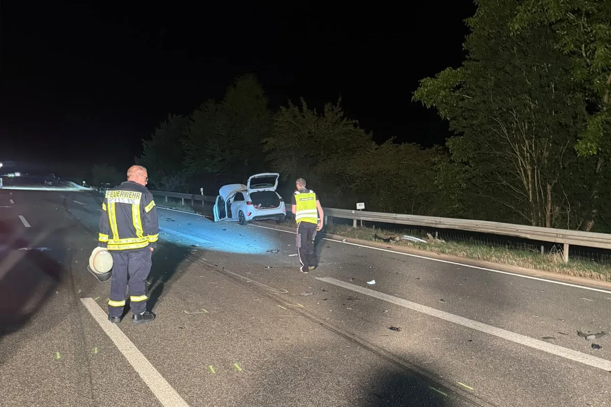 Ein beschädigtes weißes Auto steht mit geöffneter Heckklappe auf der Autobahn. Zwei Feuerwehrleute untersuchen die Unfallstelle bei Nacht.