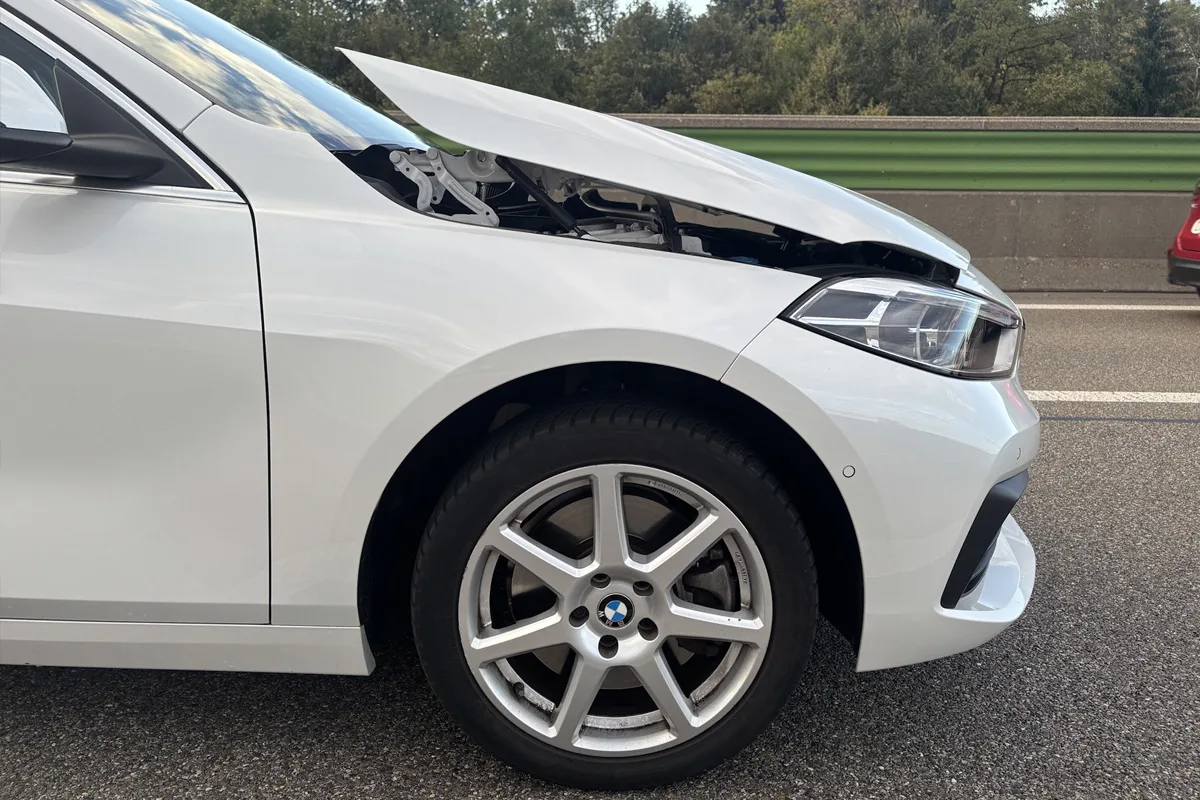 Ein weißer BMW steht am Straßenrand mit sichtbar geöffneter Motorhaube. Im Hintergrund sind Bäume und ein weiteres Fahrzeug zu sehen.