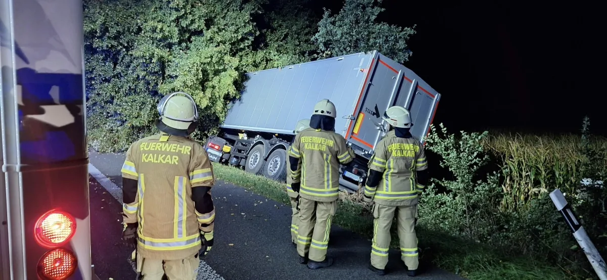 Drei Feuerwehrleute in Schutzkleidung der Feuerwehr Kalkar stehen auf einer Landstraße und betrachten einen LKW, der im Graben neben der Straße steckt. Der Unfall ereignete sich bei Nacht, was durch die künstliche Beleuchtung im Bild bestätigt wird.