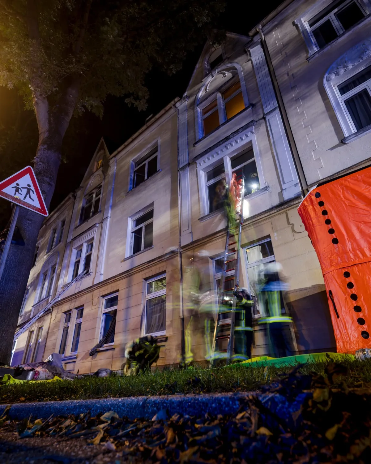 Feuerwehrleute arbeiten nachts an einem mehrstöckigen Haus. Eine Leiter ist am Gebäude aufgestellt.