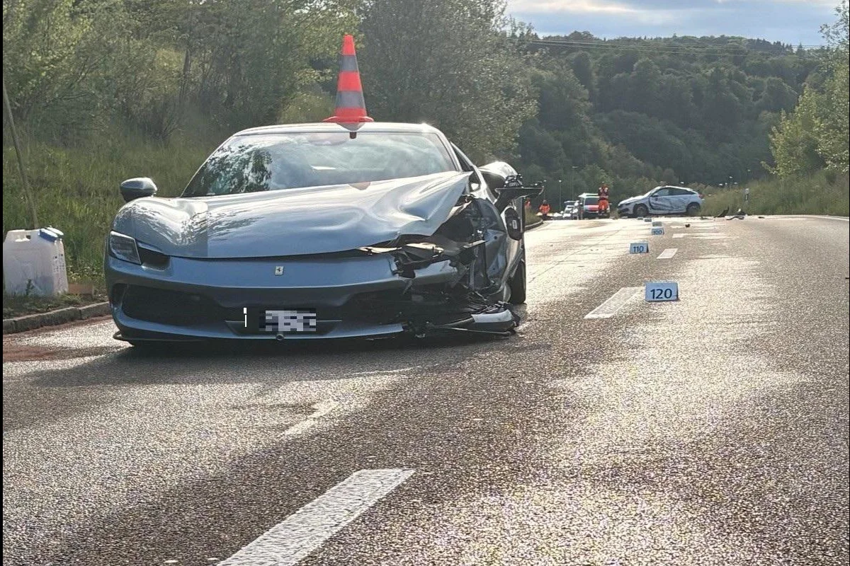 Ein stark beschädigtes Auto steht auf einer Landstraße. Ein Verkehrshut ist auf der Motorhaube platziert, und im Hintergrund sind Absperrungen und Personen sichtbar.