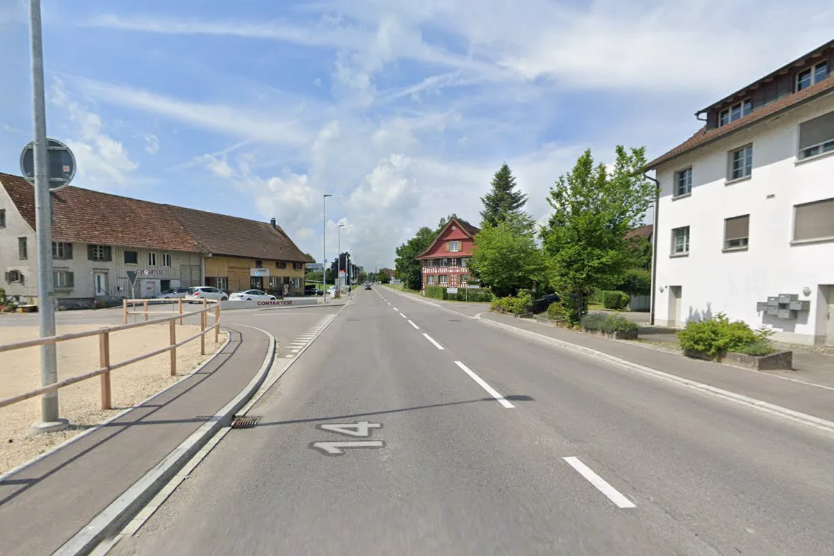 Eine ländliche Hauptstraße verläuft geradlinig, umgeben von Häusern und Bäumen unter einem klaren Himmel.