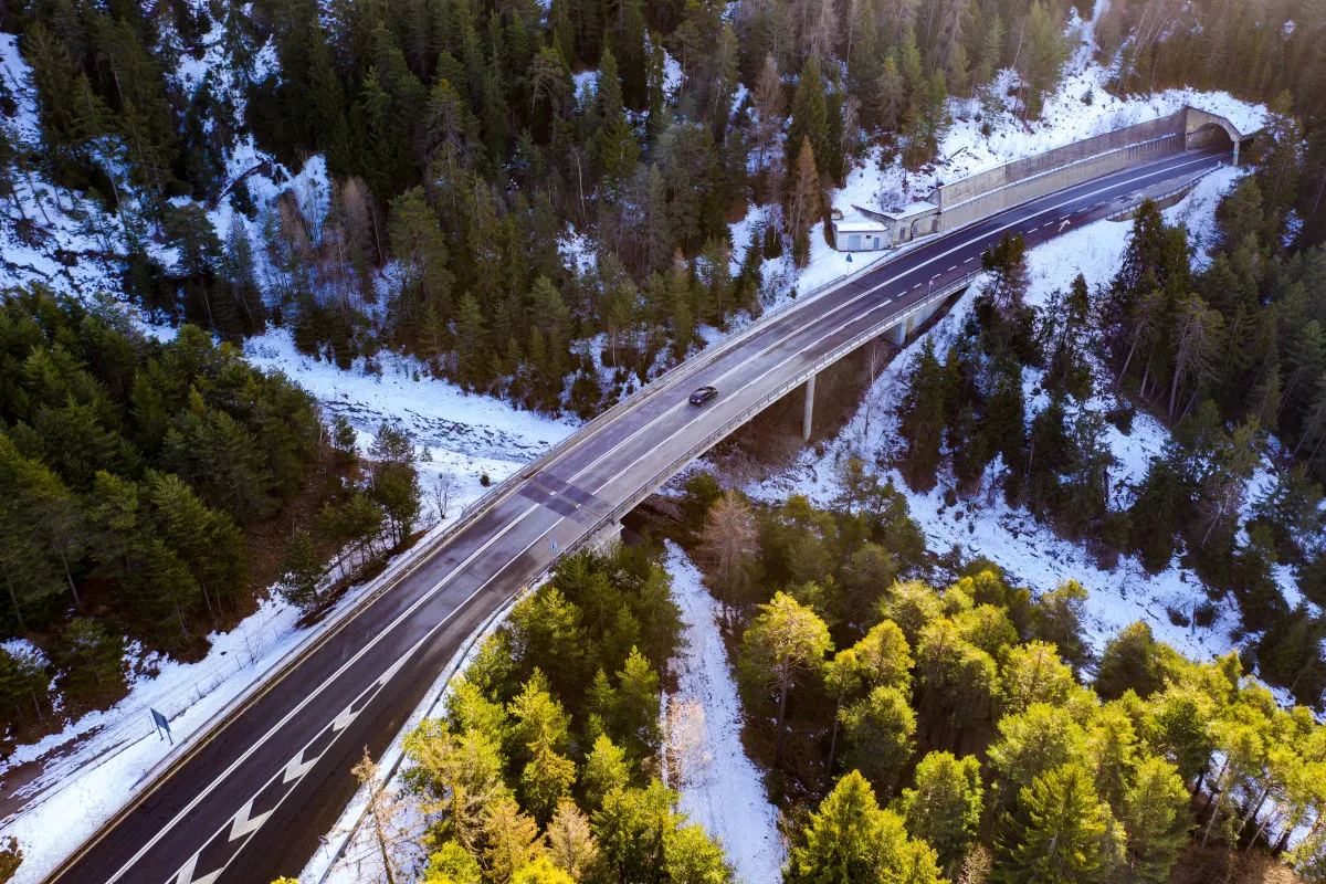 Eine Straße führt über eine Brücke, die ein winterliches Tal in einer bewaldeten Bergregion überspannt. Ein einzelnes Fahrzeug fährt darauf, während die Umgebung teils von Schnee bedeckt ist.
