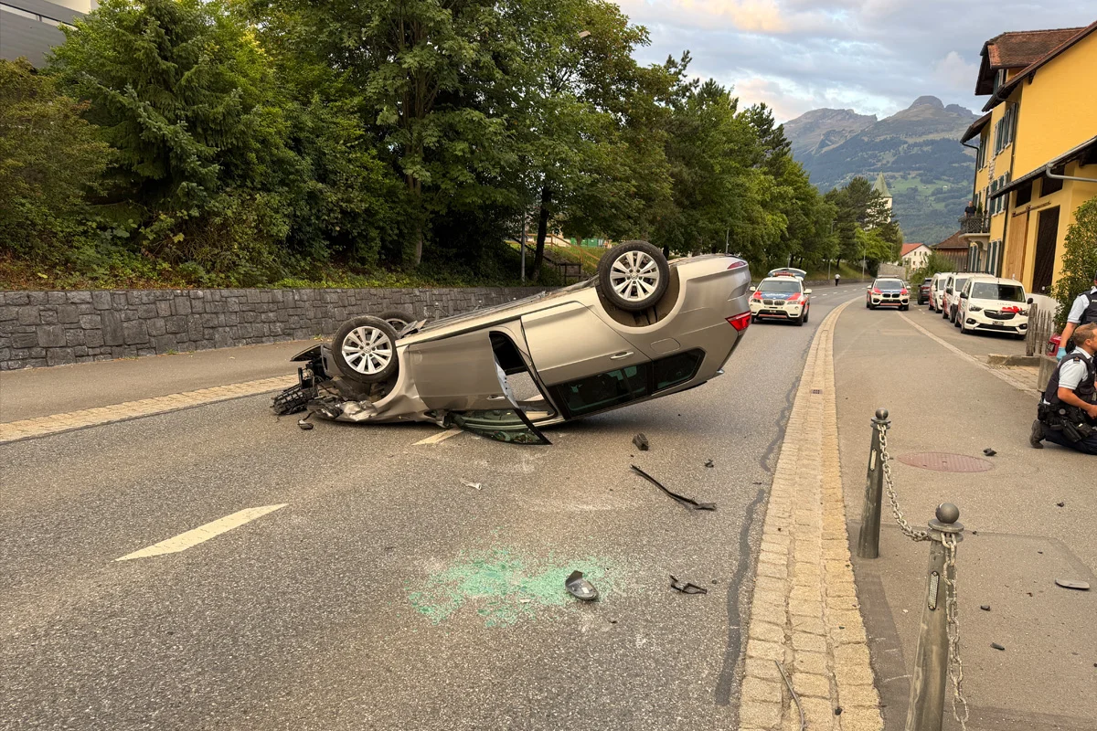 Ein umgestürztes Auto liegt auf dem Dach auf einer Straße, umgeben von Trümmern. Im Hintergrund sind mehrere Polizeifahrzeuge zu sehen.