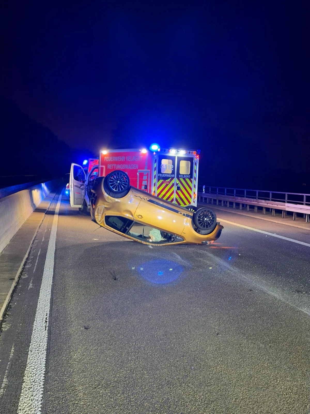 Ein Auto liegt auf dem Dach auf einer Autobahn bei Nacht. Ein Rettungswagen der Feuerwehr Velbert steht mit offenen Türen daneben.