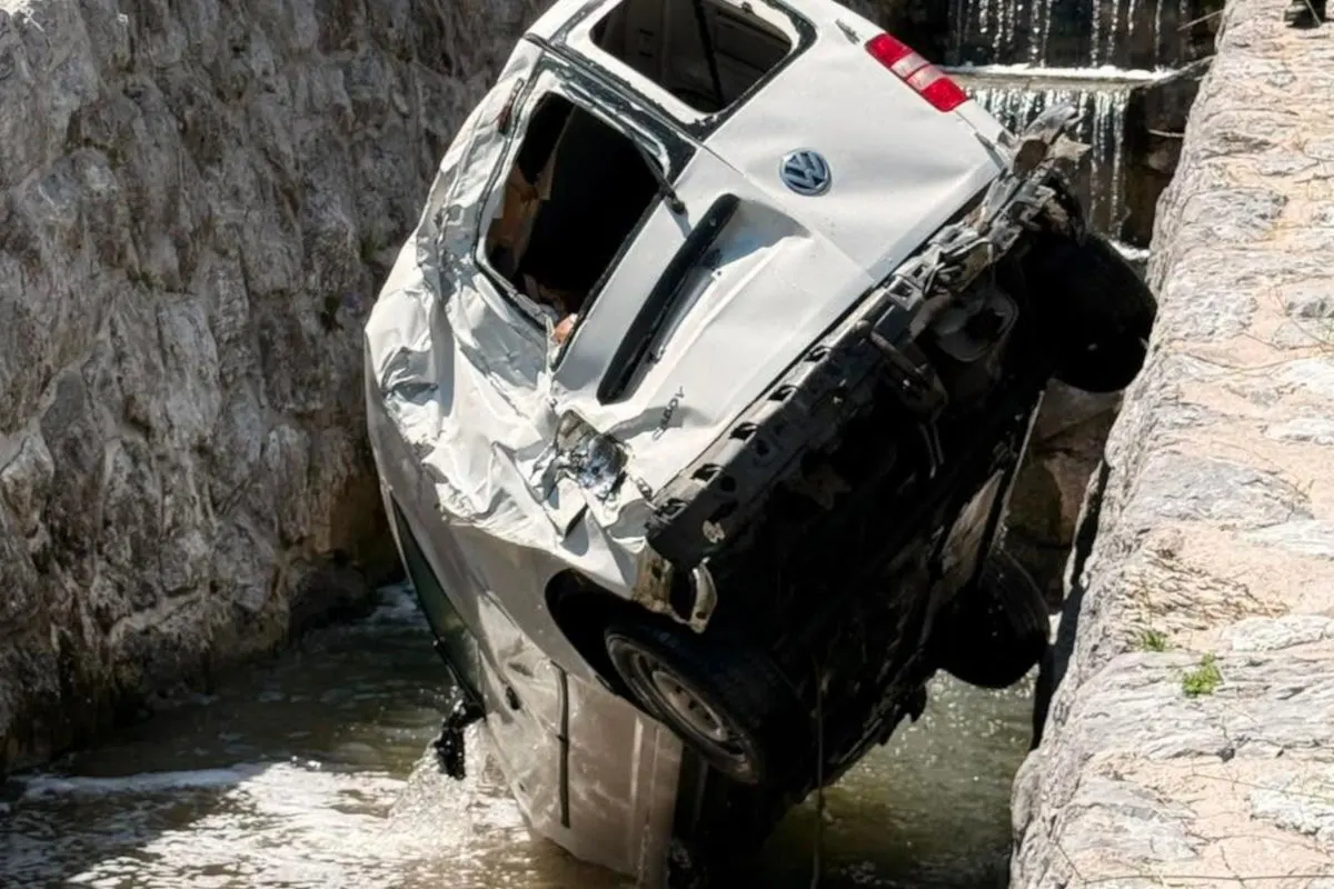 Ein weißer, stark beschädigter VW liegt schräg in einem engen Wassergraben zwischen Steinmauern. Das Auto ist verbeult und eingedrückt.