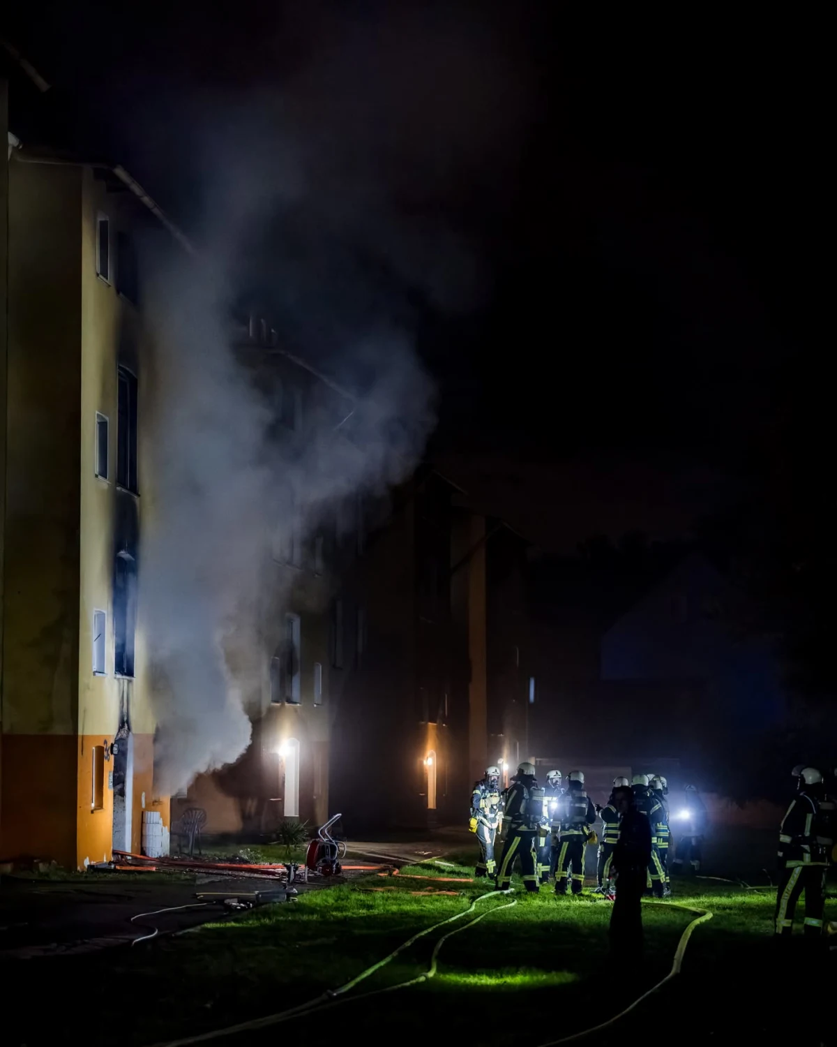 Mehrere Feuerwehrleute stehen bei Nacht vor einem verrauchten Wohngebäude. Rauch quillt aus einem Fenster im Erdgeschoss.