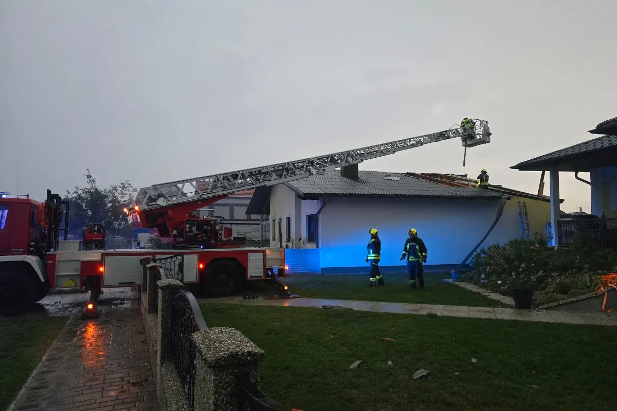 Feuerwehrleute sichern bei Abenddämmerung ein beschädigtes Hausdach mithilfe einer Drehleiter. Mehrere Einsatzkräfte sind zu sehen.