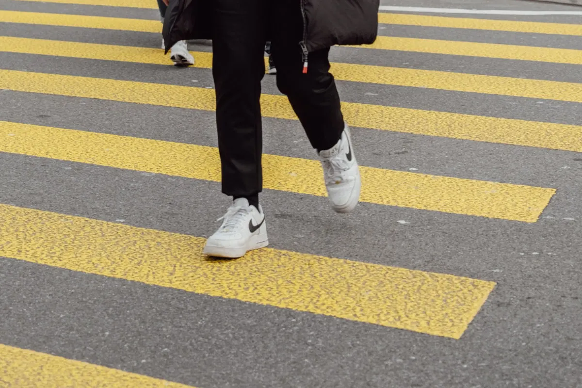 Eine Person überquert einen gelben Zebrastreifen, trägt schwarze Hosen und weiße Turnschuhe. Sichtbar ist nur der untere Teil des Körpers.