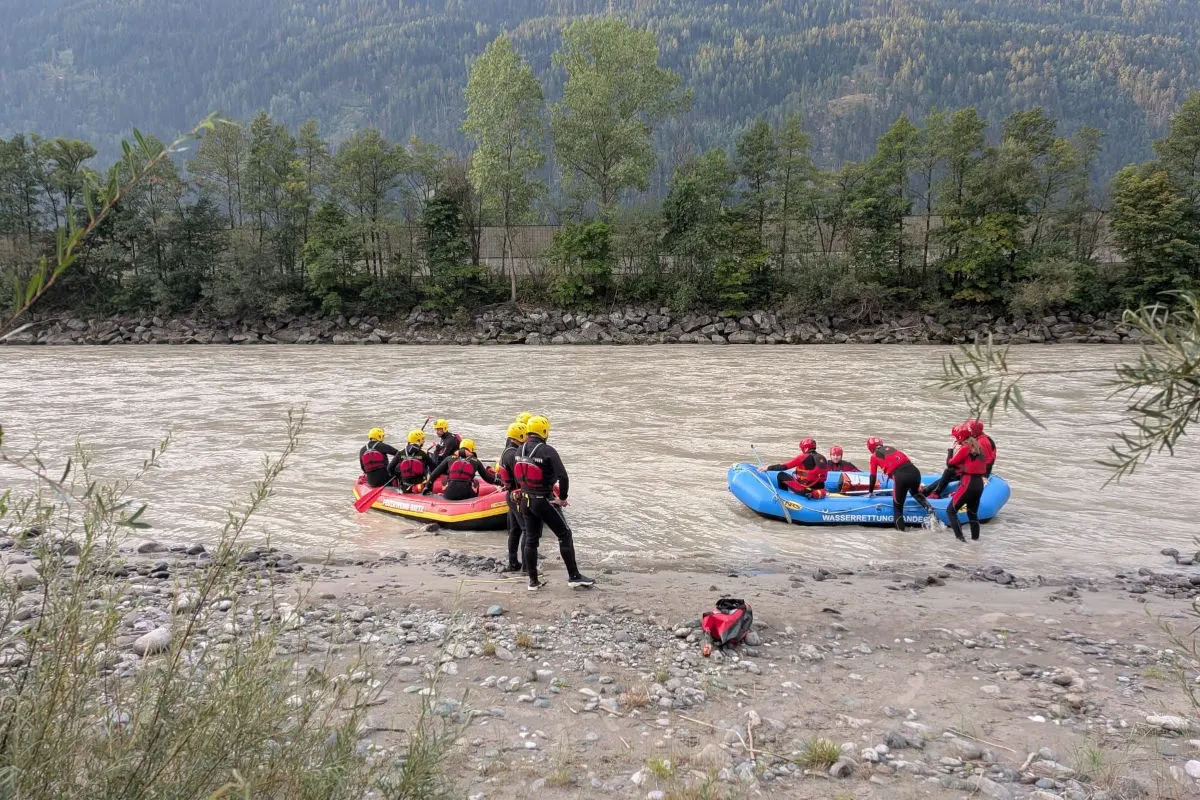 Mehrere Personen mit Helmen und Westen befinden sich in zwei Schlauchbooten auf einem Fluss nahe einem kiesigen Ufer. Im Hintergrund sind Bäume und Berge sichtbar.