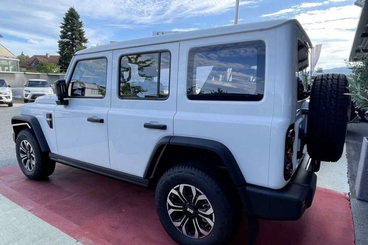 Ein moderner, weißer Geländewagen parkt auf einem rot gepflasterten Parkplatz. Im Hintergrund sind weitere Autos und ein blauer Himmel erkennbar.