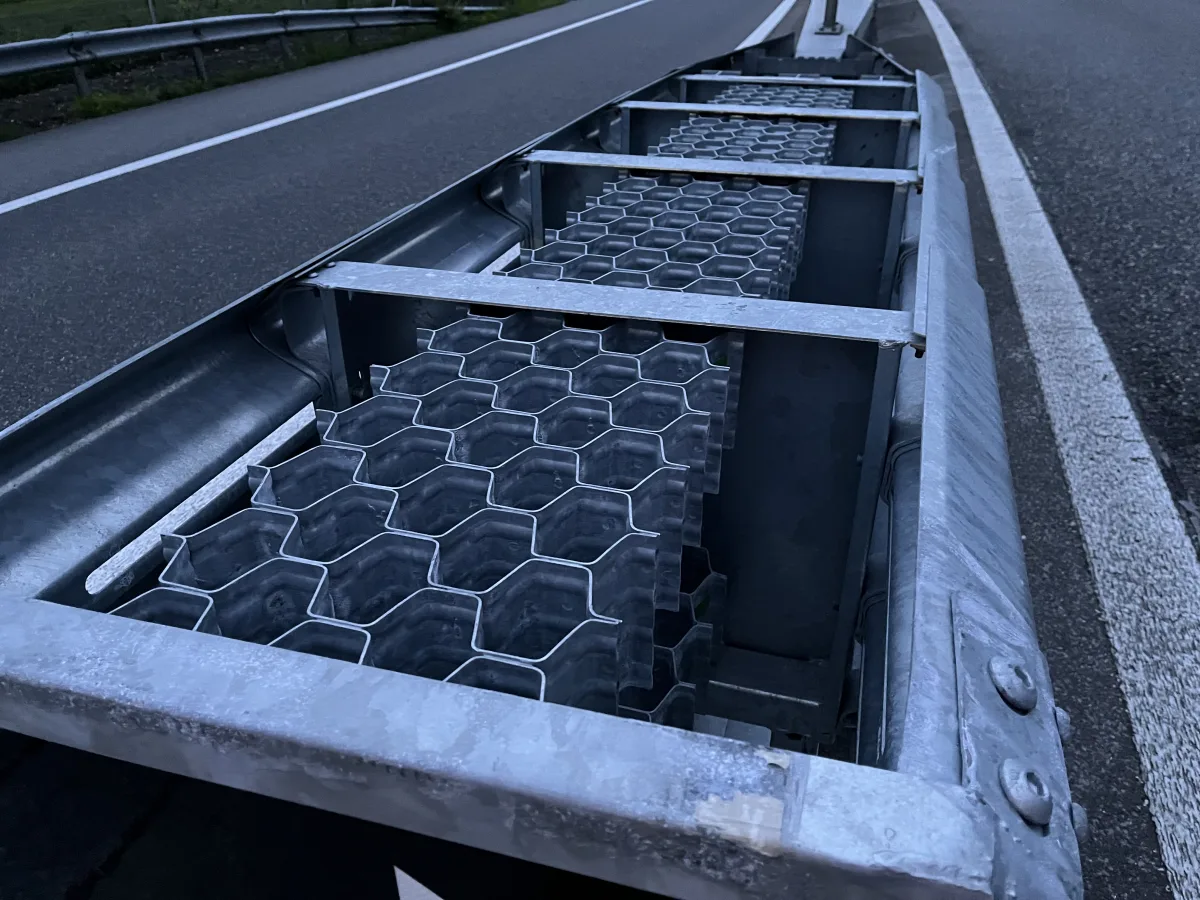 Eine Nahaufnahme zeigt eine beschädigte Leitplanke mit einer wabenförmigen Struktur. Die Szene spielt sich bei Dämmerung auf einer Straße ab.