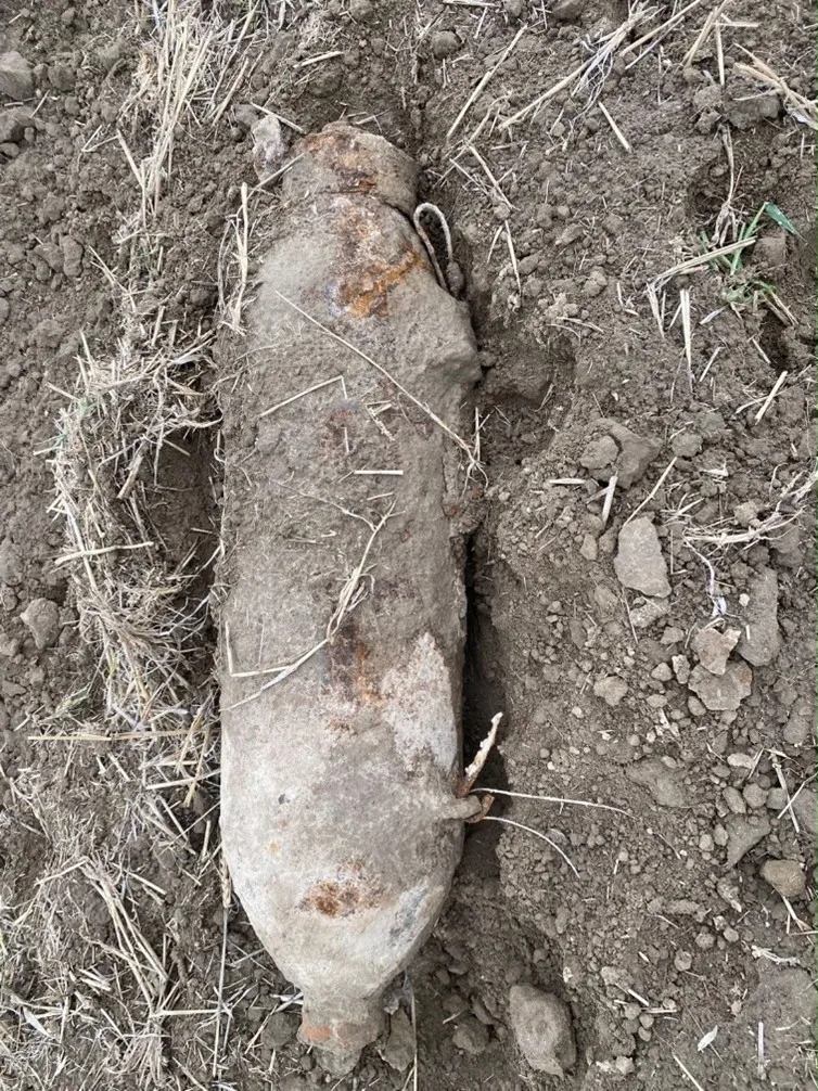 Im Bild liegt eine verrostete Fliegerbombe zur Hälfte aus dem Erdreich ausgegraben. Das Gelände besteht aus trockener, grasbewachsener Erde.