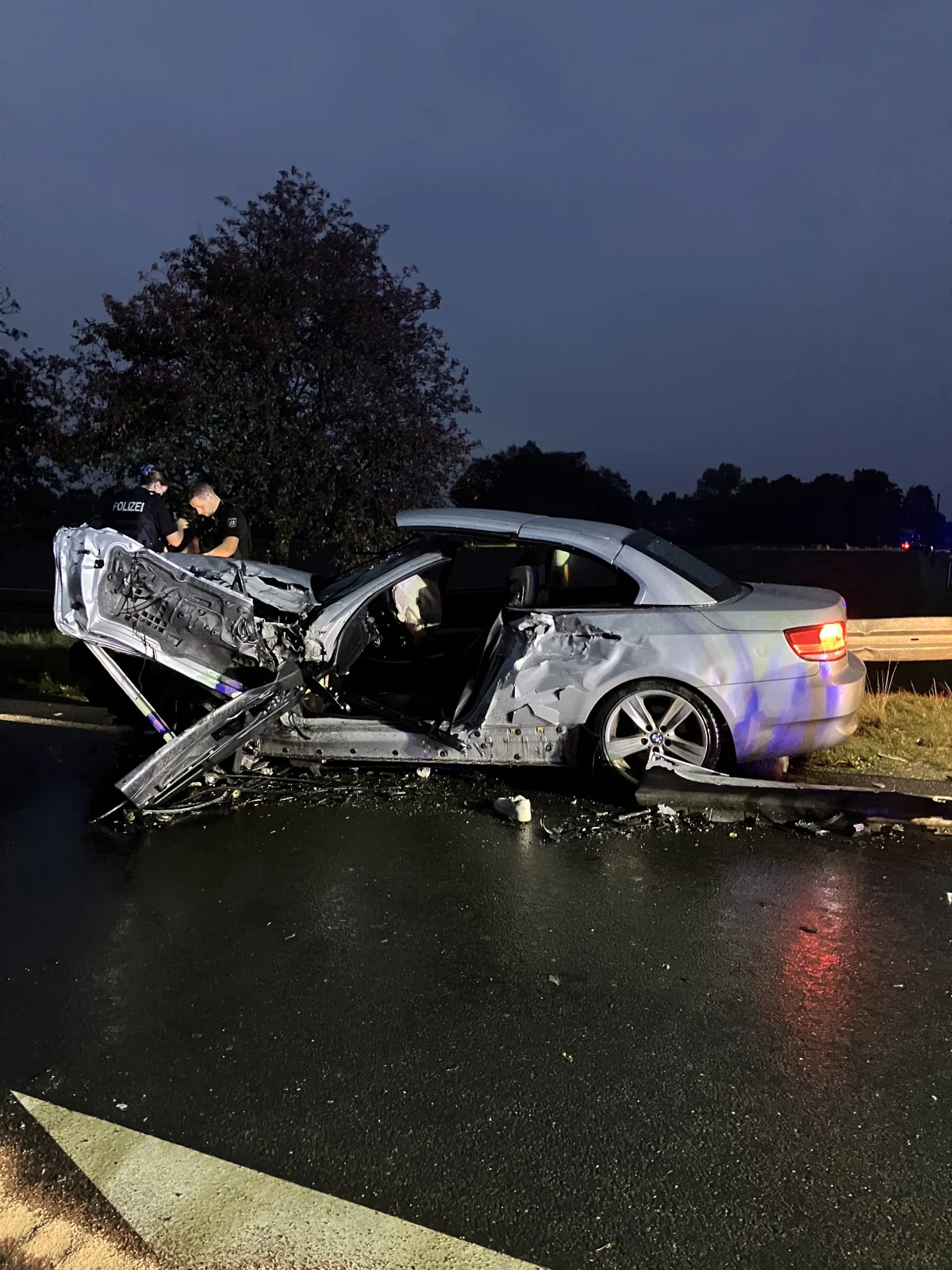 Ein stark beschädigtes silbernes Auto steht mit geöffneten Türen auf der Straße. Zwei Polizisten sind daneben im Einsatz.