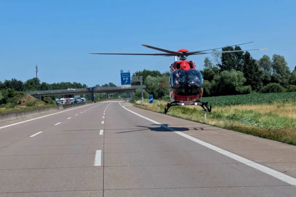 Ein roter Rettungshubschrauber befindet sich im Landeanflug auf einer leeren Autobahn. Im Hintergrund sind Bäume und ein Überführungsbauwerk zu sehen.