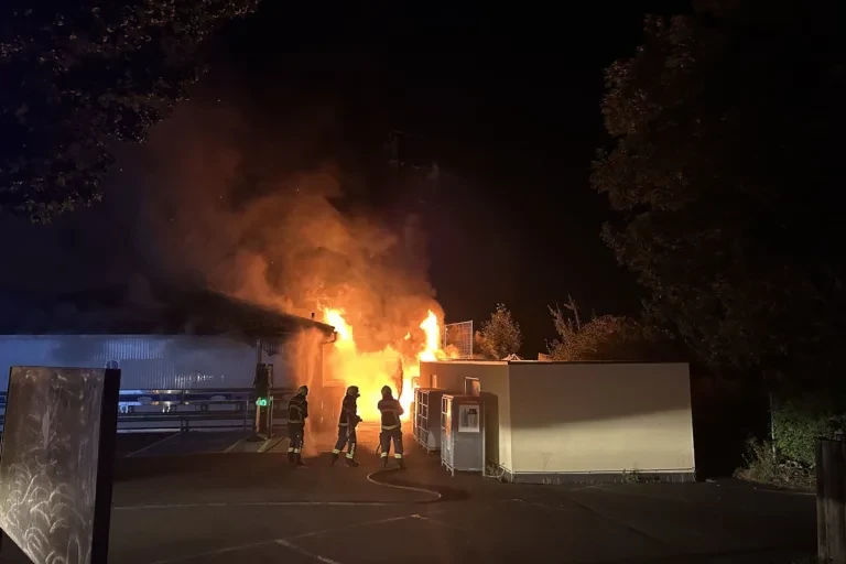 Feuerwehrleute stehen nachts vor einem brennenden Gebäude, Flammen schlagen aus dem Dach.