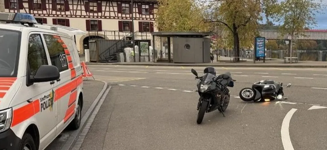 Ein Polizeifahrzeug steht links neben einem umgestürzten Motorrad auf einer leeren Straße. Im Hintergrund sind Bäume und Gebäude zu sehen.