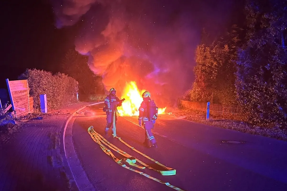 Auf der Straße sind zwei Feuerwehrleute zu sehen, die sich einem großen Feuer nähern. Es ist nachts, und der Himmel ist dunkel.