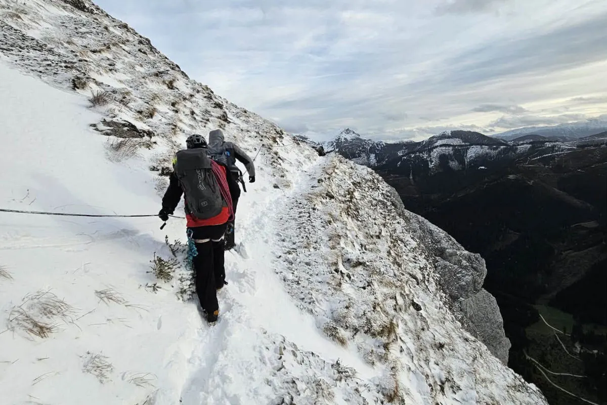 Zwei Personen sind auf einem schneebedeckten und schmalen Grat in den Bergen zu sehen. Sie sind durch Seile gesichert und tragen Bergsteigerausrüstung.