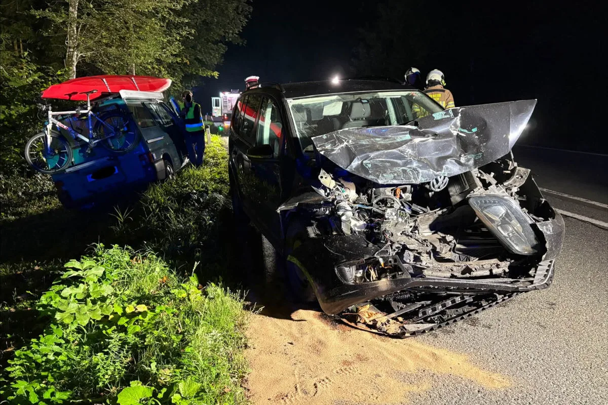 Ein schwarzer VW mit starkem Frontschaden steht auf der Straße, während ein blauer Van in der Böschung parkt und ein rotes Kanu sowie ein Fahrrad trägt. Zwei Personen in Schutzkleidung sind ebenfalls zu sehen.