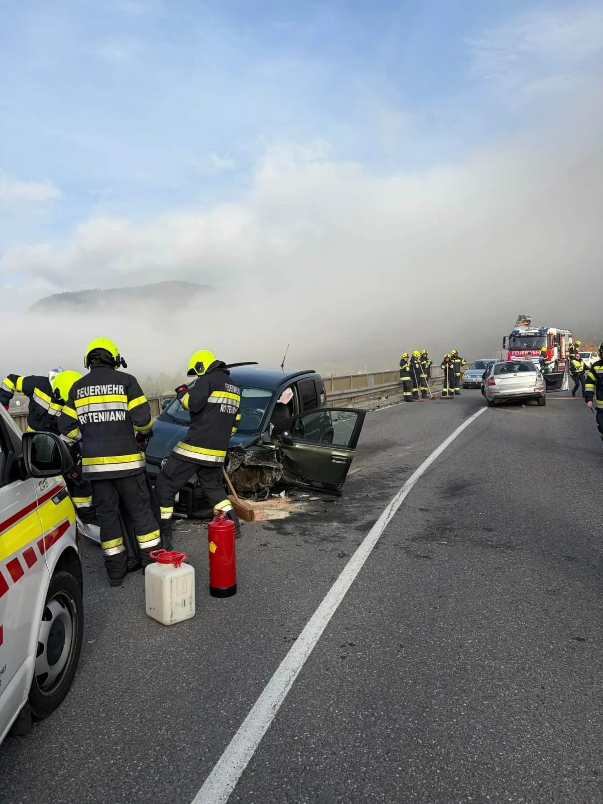 Mehrere Feuerwehrleute mit gelben Helmen arbeiten an einem beschädigten Auto auf einer Straße. Ein weiteres Feuerwehrfahrzeug ist in der Nähe sichtbar.