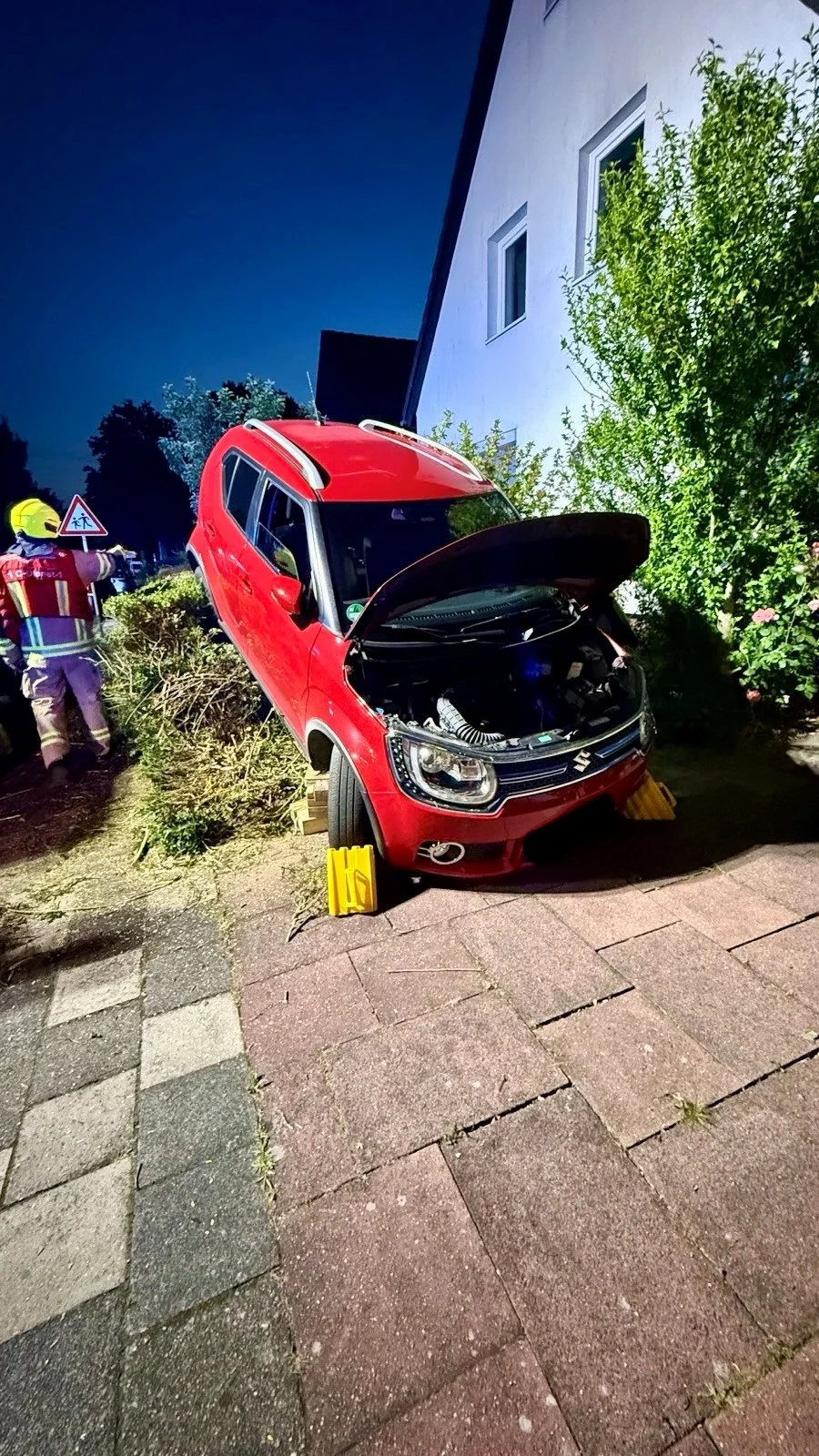 Ein rotes Auto, dessen Motorhaube geöffnet ist, hängt vorne an einem Bordstein fest. Daneben ist eine Person in Rettungskleidung zu sehen.