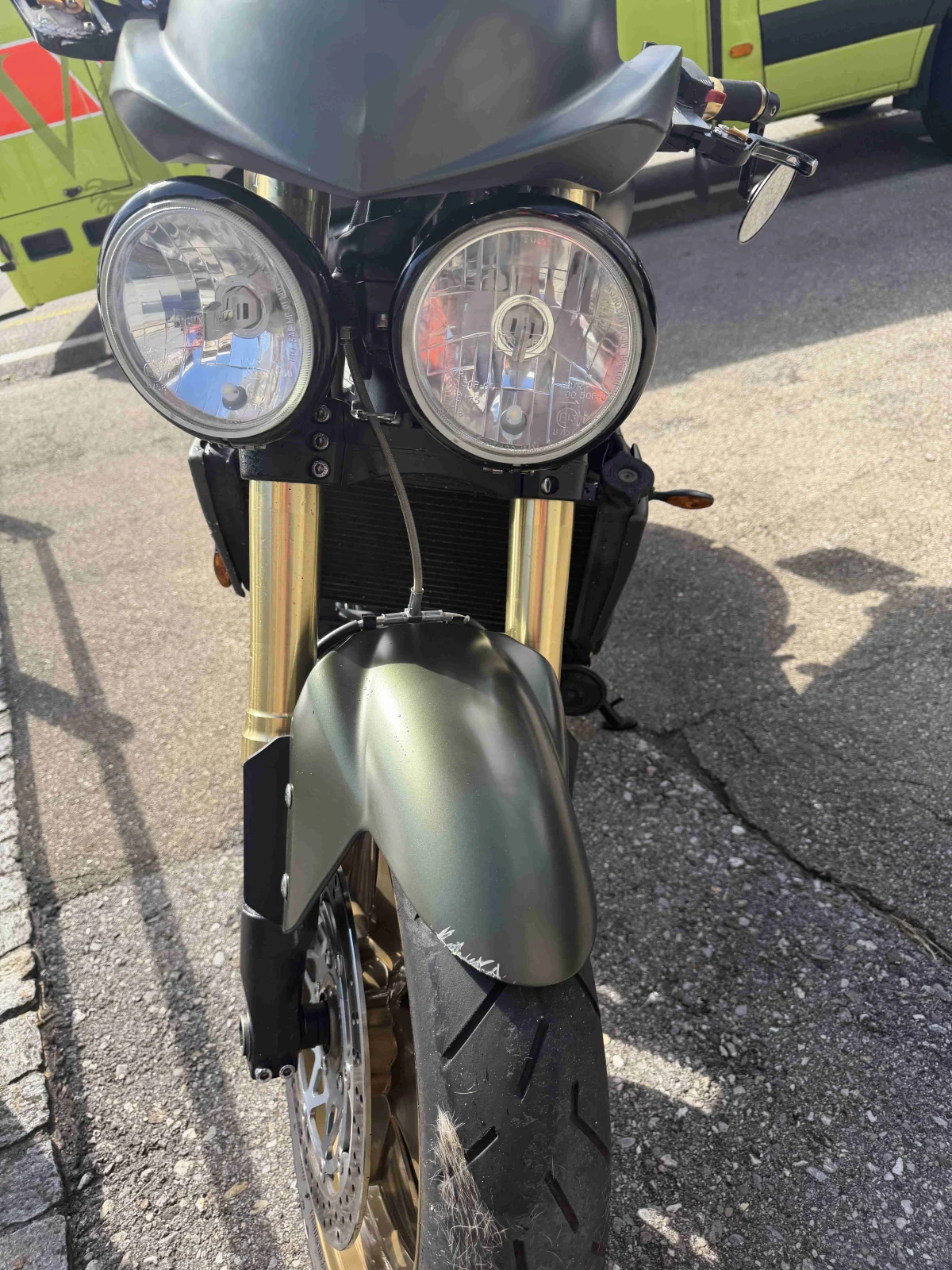 Ein Motorrad steht frontal im Bild, der Kotflügel zeigt leichte Kratzer. Im Hintergrund ist ein gelb-grünes Fahrzeug sichtbar.