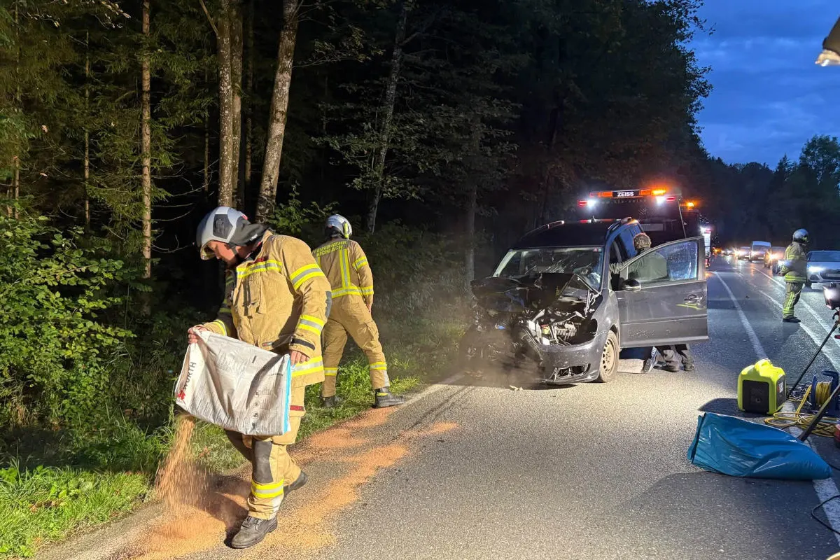 Ein Feuerwehrmann in Schutzkleidung streut Bindemittel neben einem beschädigten Auto auf einer Landstraße in der Nacht. Ein weiteres Feuerwehrfahrzeug und andere Einsatzkräfte sind im Hintergrund zu sehen.