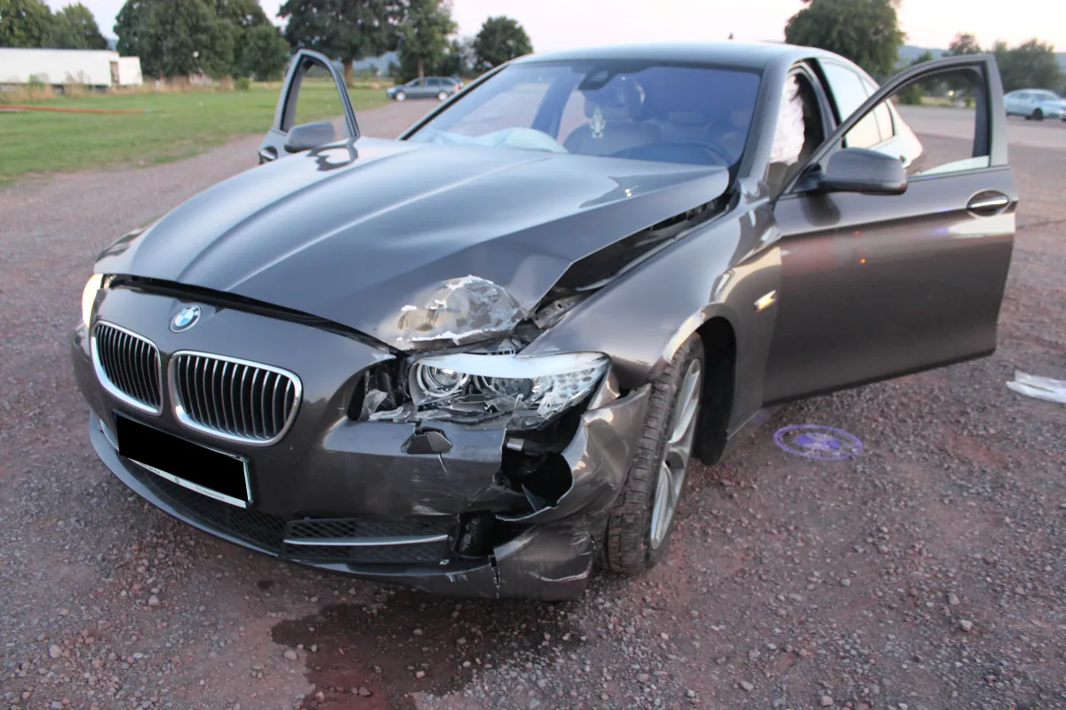 Ein schwarzer BMW mit deutlichen Frontschäden und geöffneter Motorhaube steht auf einem Schotterplatz. Die Türen sind geöffnet und die Airbags ausgelöst.