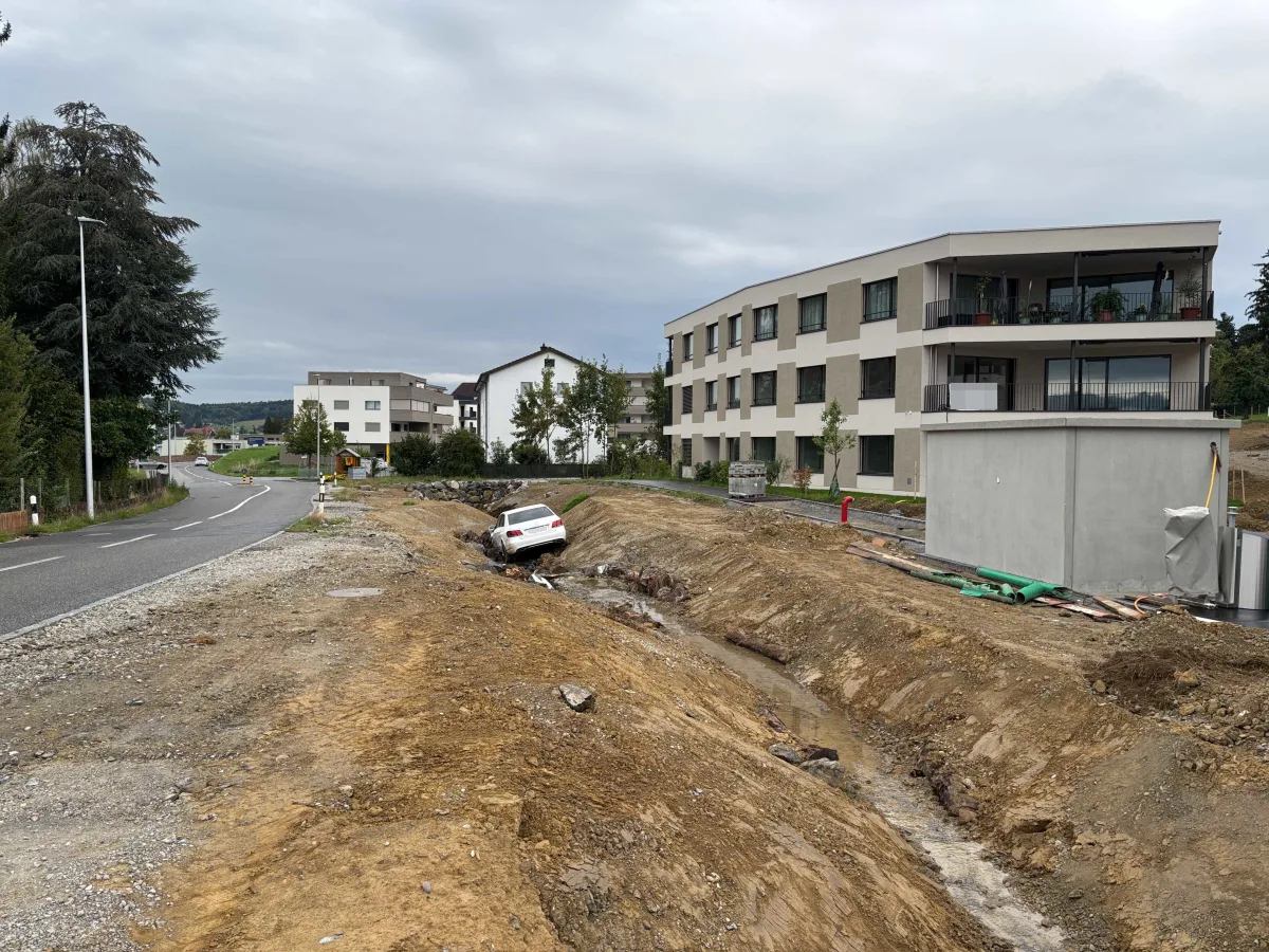 Ein weißes Auto ist in einen Graben neben einer Baustelle gerutscht. Im Hintergrund sind eine Straße und moderne Wohngebäude zu sehen.