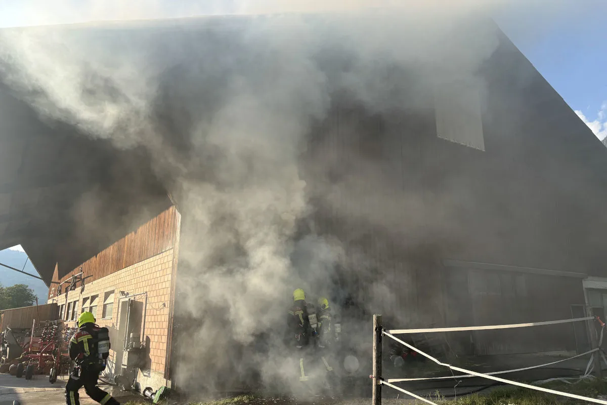 Mehrere Feuerwehrleute in Schutzkleidung stehen vor einem Gebäude, das von dichtem Rauch umgeben ist. Das Gebäude sieht landwirtschaftlich aus.