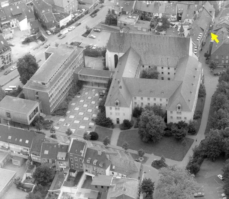 Eine Schwarz-Weiß-Luftaufnahme eines großen Gebäudekomplexes in einem städtischen Gebiet, umgeben von Straßen und Bäumen.