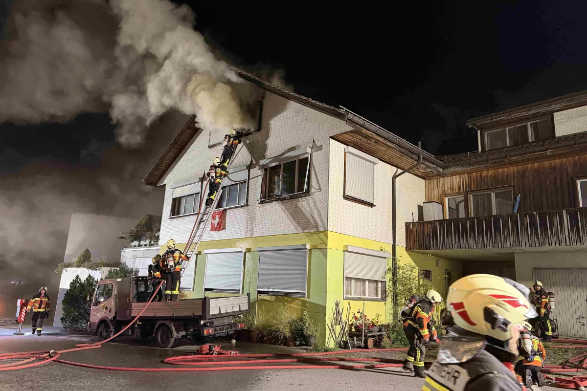 Mehrere Feuerwehrleute arbeiten an einem brennenden Wohnhaus bei Nacht. Rauch steigt vom Dach auf.