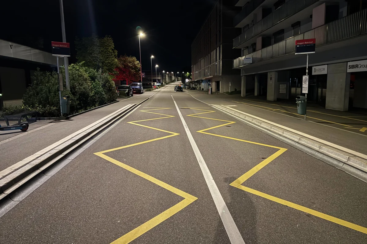 Eine leere Straße bei Nacht, erleuchtet von Straßenlaternen. Gelbe Markierungen sind auf der Fahrbahn sichtbar, flankiert von Gebäuden.