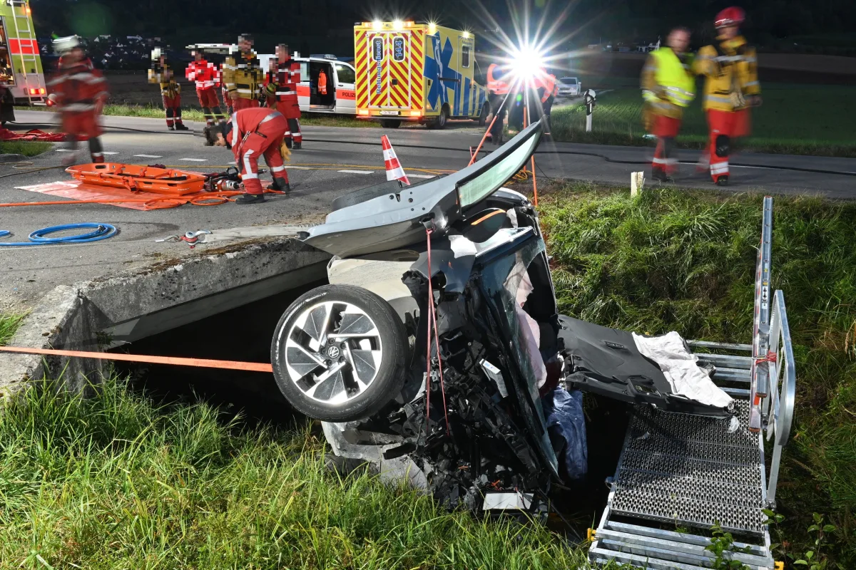 Ein Auto liegt auf dem Dach in einem Straßengraben. Im Hintergrund sind mehrere Rettungskräfte und ein Krankenwagen zu sehen.